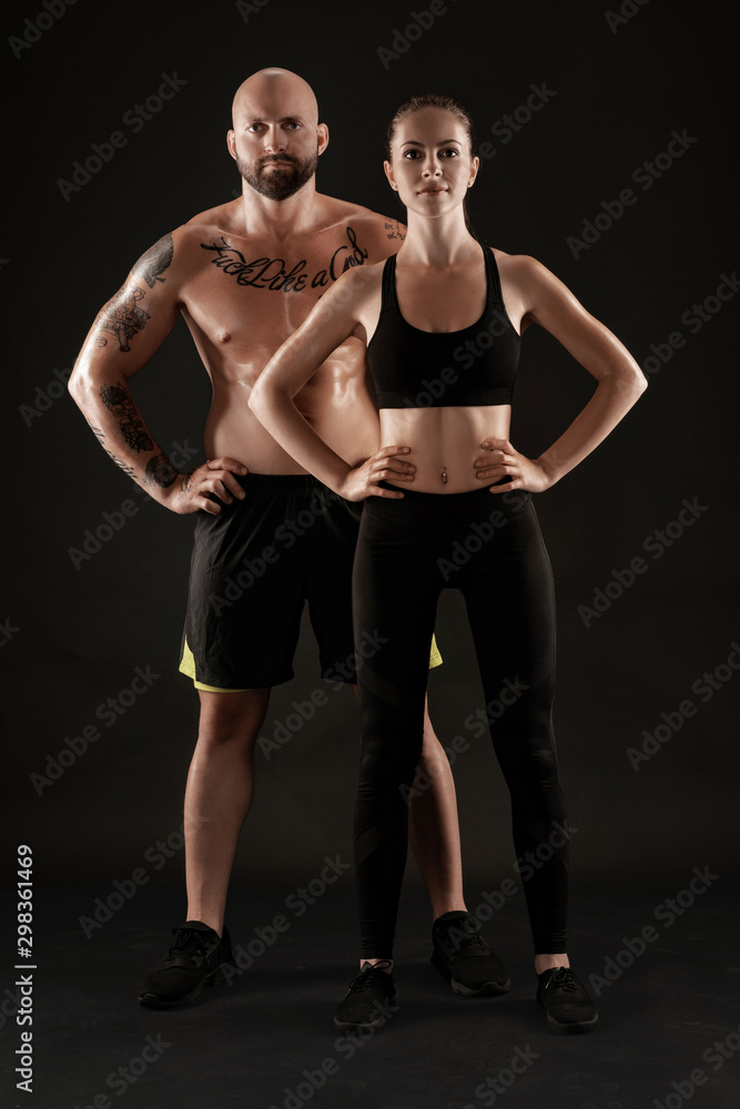 Athletic man in shorts and sneakers with brunette woman in leggings and top posing on black background. Fitness couple, gym concept. Athletic man in shorts and sneakers with brunette woman in leggings and top posing on black background. Fitness couple, gym concept.