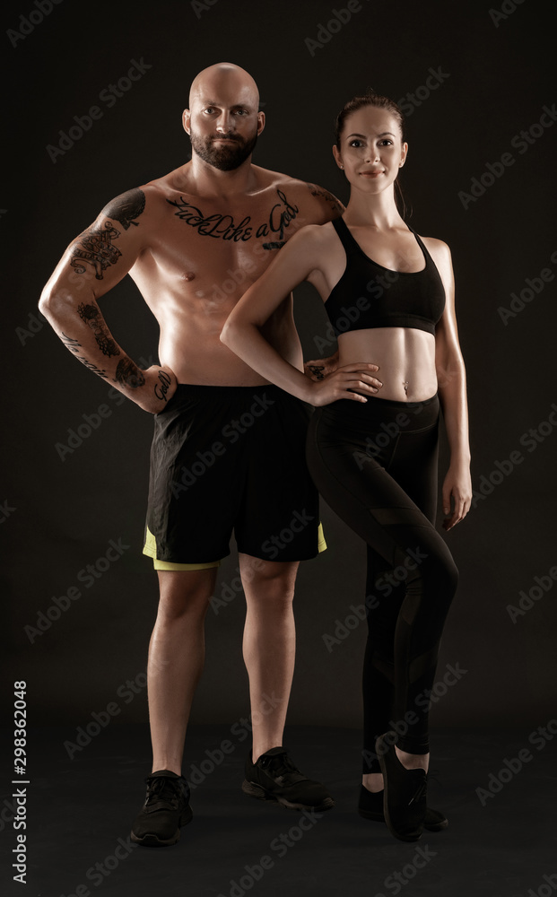 Athletic man in shorts and sneakers with brunette woman in leggings and top posing on black background. Fitness couple, gym concept.