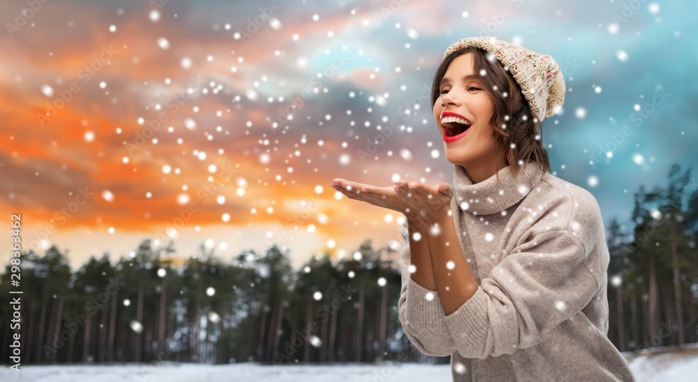 christmas, season and people concept - happy smiling young woman in knitted hat and sweater sending air kiss over winter forest and snow background christmas, season and people concept - happy smiling young woman in knitted hat and sweater sending air kiss over winter forest and snow background