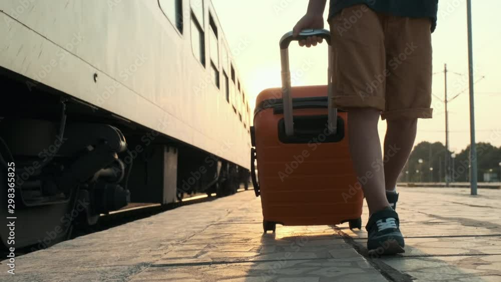 Happy boy walks along the platform of the train station and rolls an orange suitcase behind him in the rays of a bright sunny sunset slow motion. Child goes vacation by train. Tourism, travel Happy boy walks along the platform of the train station and rolls an orange suitcase behind him in the rays of a bright sunny sunset slow motion. Child goes vacation by train. Tourism, travel