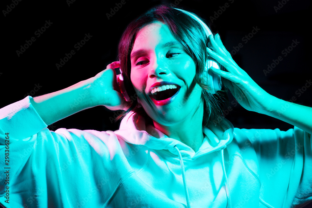 nightlife, technology and people concept - happy young woman in headphones wearing hoodie listening to music and dancing in neon lights over black background nightlife, technology and people concept - happy young woman in headphones wearing hoodie listening to music and dancing in neon lights over black background