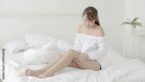 Beautiful young asian woman sitting on a bed stroking legs with soft smooth skin in the bedroom, girl applying body cream and lotion with treatment care, healthy and wellness concept.