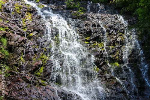 Close up of a powerful mountain waterfall cascading down the rocky  red cliff covered by green and golden sunlit moss