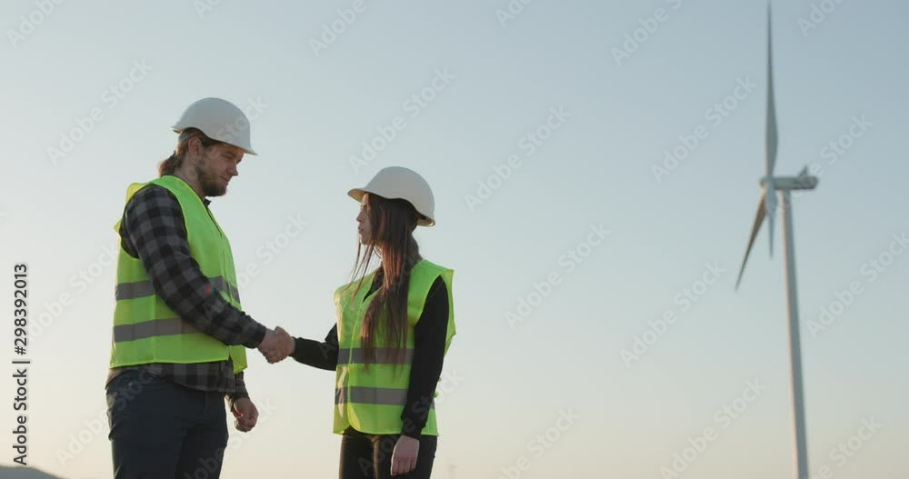 Engineers man and woman in helmets shaking hands on the windmill turbine background. Engineers man and woman in helmets shaking hands on the windmill turbine background.