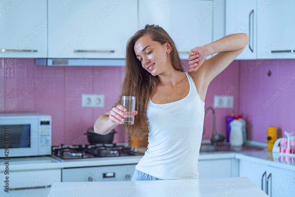 Happy sleepy woman stretching and drinks a glass of clean purified morning water in early morning after waking up in the kitchen at home. Healthy lifestyle. Beginning and start of a new good day