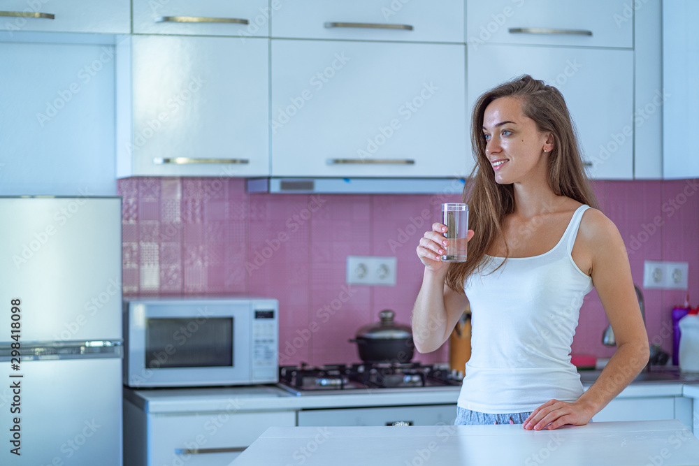 Happy sleepy joyful woman drinks a glass of clean purified morning water in early morning after waking up in the kitchen at home. Healthy lifestyle. Beginning and start of a new good day Happy sleepy joyful woman drinks a glass of clean purified morning water in early morning after waking up in the kitchen at home. Healthy lifestyle. Beginning and start of a new good day