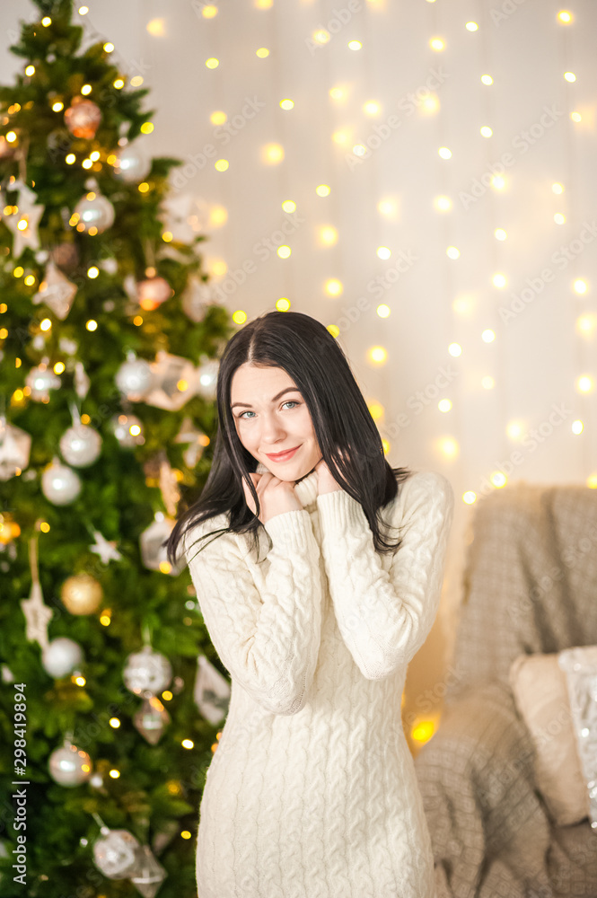 Lovely girl in the morning in a white knitted dress in the New Year’s interior. Woman at home at Christmas time in a living room in a rustic style near a New Year tree. Lovely girl in the morning in a white knitted dress in the New Year’s interior. Woman at home at Christmas time in a living room in a rustic style near a New Year tree.