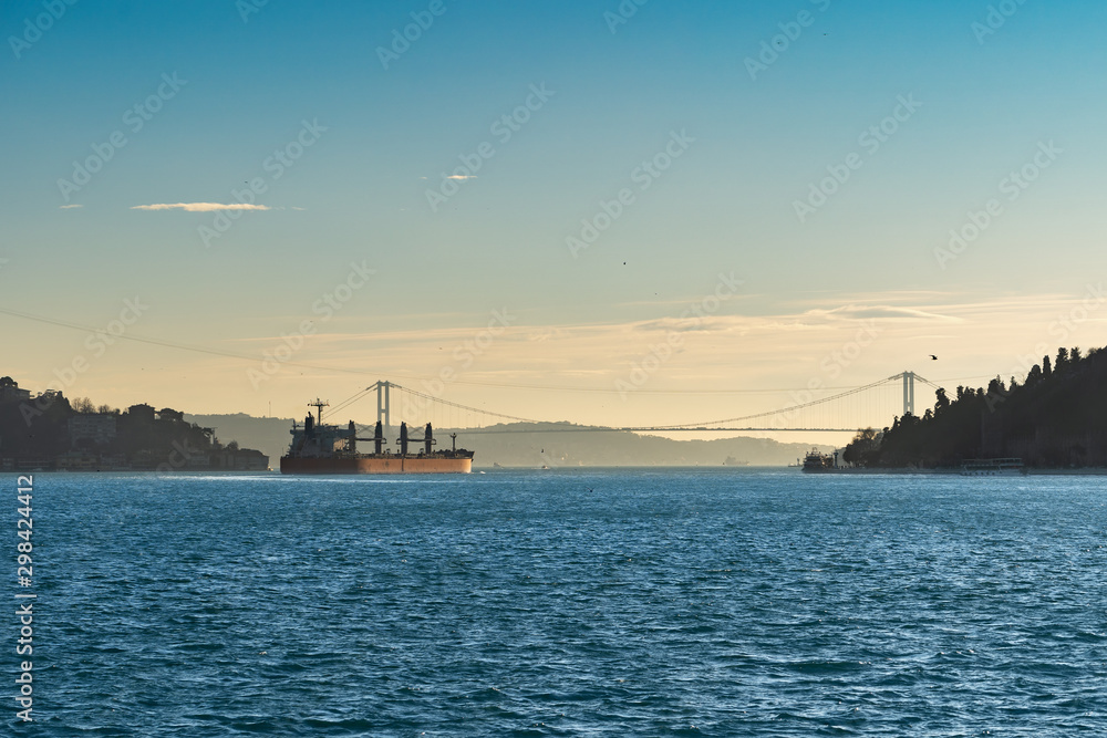 A large cargo ship - bulk-carrier, sailing along the blue waves of the strait, in the light of the evening sun, is approaching a bridge over the Bosphorus Strait.