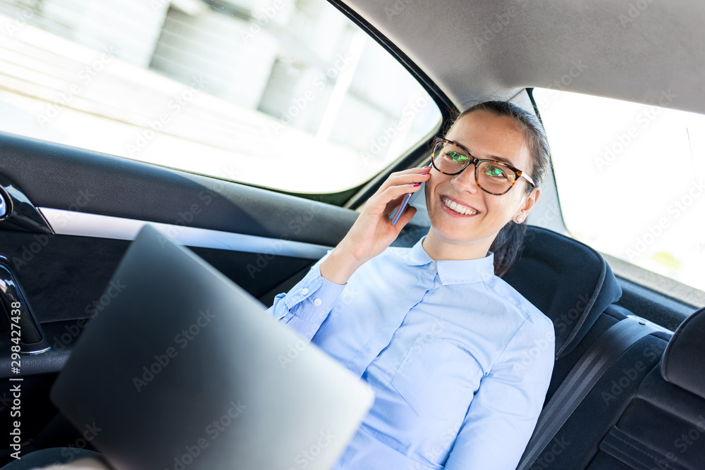 Young businesswoman talking on the phone in the back seat of the car and holding laptop.