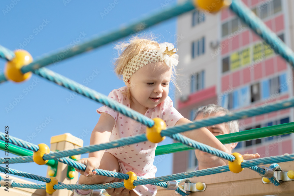 Active little child playing on climbing net and jumping on trampoline at school yard playground. Kids play and climb outdoors on sunny summer day. Cute girl on nest swing at preschool sport center.