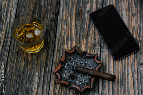 Saturated, contrasting photo of a glass of whiskey with ice, smoldering cigar on an ashtray and a smartphone on an old textured wooden table. View from above	