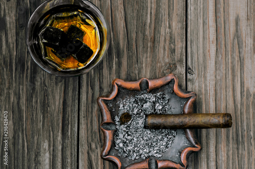 Saturated, contrasting photo of a glass of whiskey with whisky stones and a smoldering cigar on an ashtray on an old textured wooden table. View from above