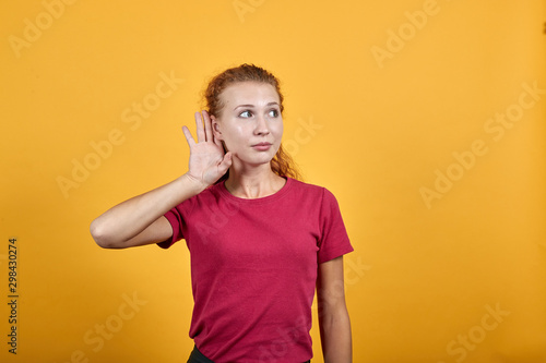 Young European woman as if listening to something by putting her right hand on ear. Charming lady in pinky garment with long wavy dark hair standing over isolated yellow background.