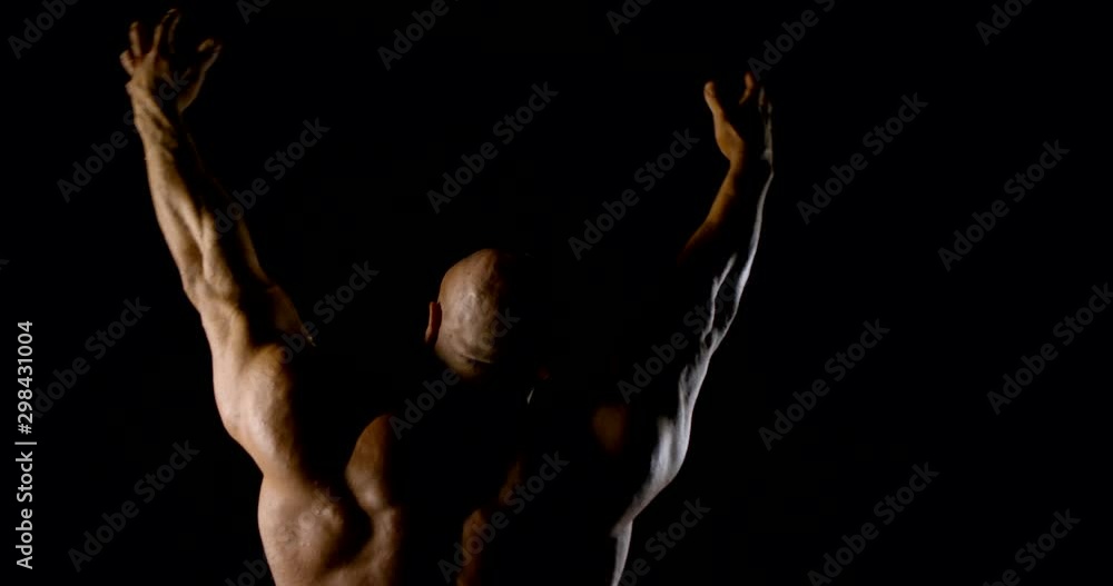 brawny man is lifting hands up and tensing muscles of back, showing them for camera in darkness brawny man is lifting hands up and tensing muscles of back, showing them for camera in darkness