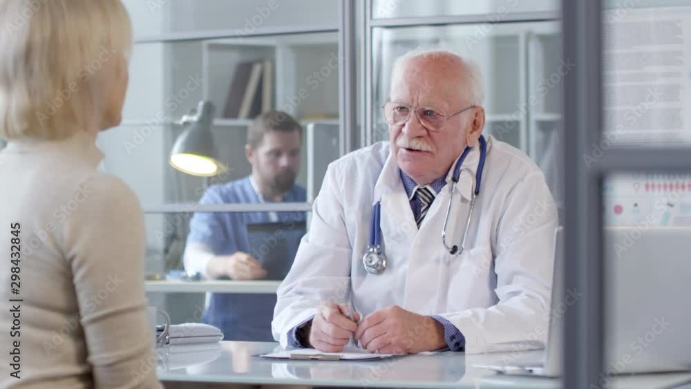 Senior doctor in lab coat sitting at desk and speaking with female patient during medical consultation in clinic; male practitioner working in background behind glass wall Senior doctor in lab coat sitting at desk and speaking with female patient during medical consultation in clinic; male practitioner working in background behind glass wall