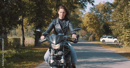 Pretty smiling European young woman driving a motorbike wearing leather jacket in forest with vibrant, colorful golden autumn leaves on sunny day. Frontal view, Slow Motion, Utrecht , Netherlands photo