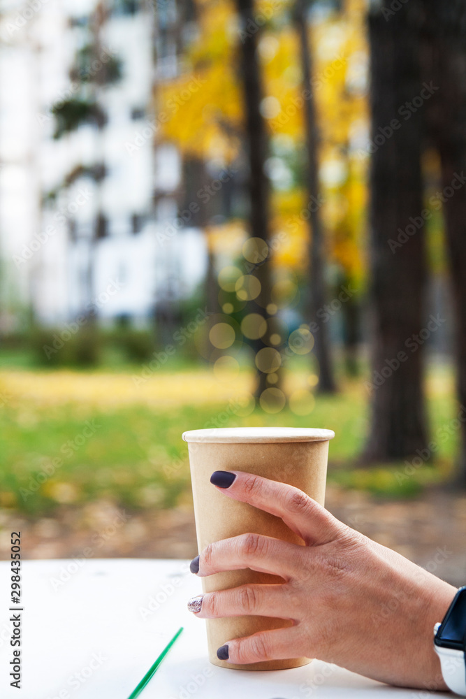 Girl's hand holds a cup of drink. A table in the autumn park. Autumn mood on a background of yellow foliage Girl's hand holds a cup of drink. A table in the autumn park. Autumn mood on a background of yellow foliage