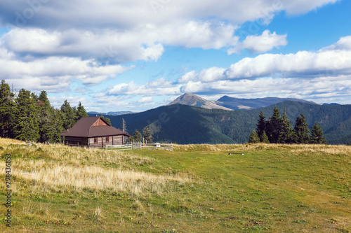 The landscape with the wooden hut and fence on the lawn with green fir trees, high mountains covered by forests, sky with clouds. Autumn sunny day.