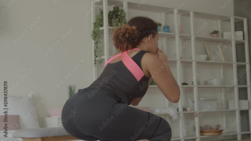 Rear view of determined sporty fit african american woman in sportswear crouching and improving muscles on lower body at home. Active fitness female working out two-leg deep squat exercise indoors. Rear view of determined sporty fit african american woman in sportswear crouching and improving muscles on lower body at home. Active fitness female working out two-leg deep squat exercise indoors.