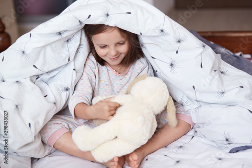 Portrait of cute girl and dog toy under blanket, little female kid and dog toy playing together games under white blanket, chirming child spending morning time in cosy room. Childhood concept. photo