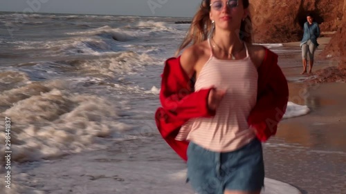 A beautiful girl with long blond hair, in a red jacket, with unusual, rectangular purple glasses, a chain, denim shorts, with bare feet, a striped T-shirt runs along the shore, the beach, wet sand photo
