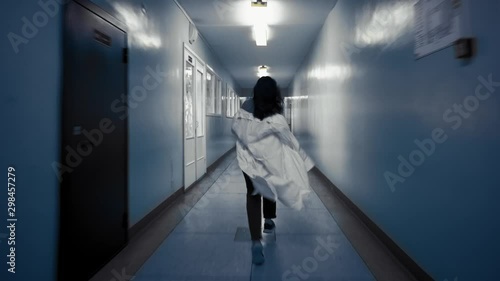 Tracking shot of zombie woman with black hair in laboratory coat walks down dark corridor goes behind door and falls,view from back. Above doorway inscription in Russian exit.