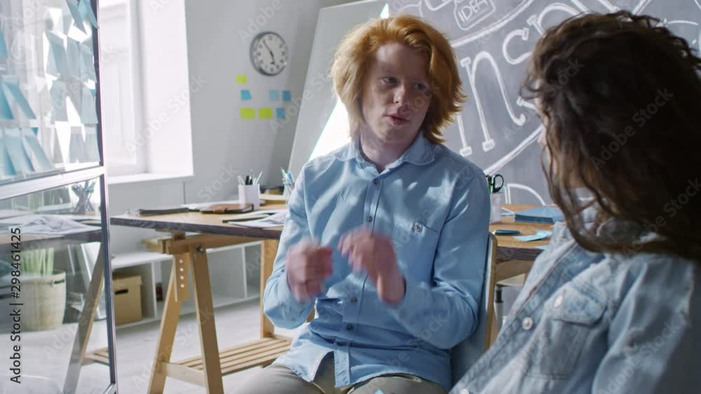 Young redhead businessman explaining something to female colleague while sitting in modern office with chalkboard wall Young redhead businessman explaining something to female colleague while sitting in modern office with chalkboard wall