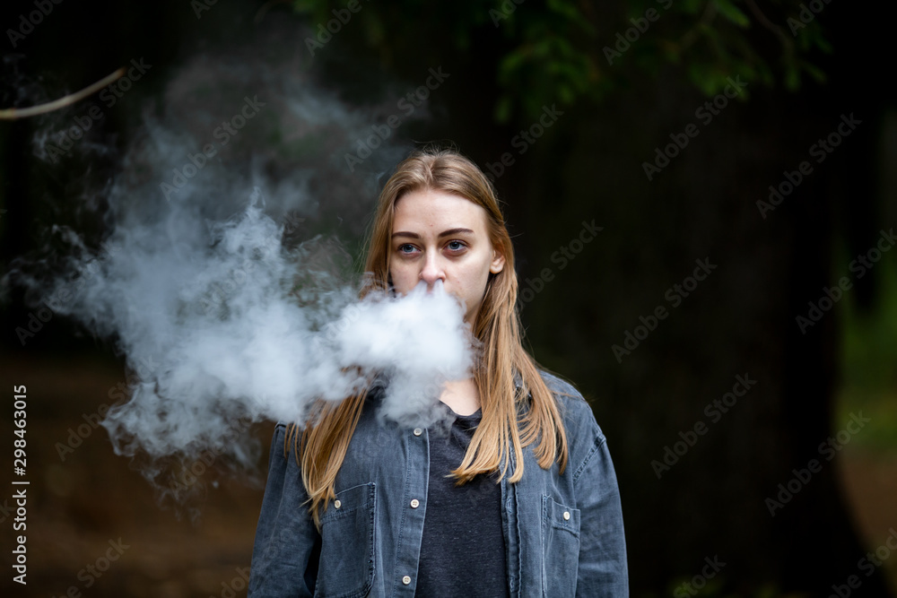 Vape teenager. Young white addict caucasian teenage girl with bruises under the eyes in casual clothes vaping electronic cigarette with cannabis liquid in the dark forest in the autumn evening.