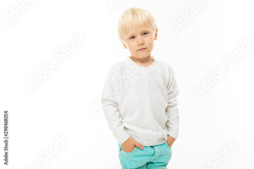 Little boy with short blonde hair, blue eyes, cute appearance, in white jacket, light blue pants, standing with his hands in his pockets
