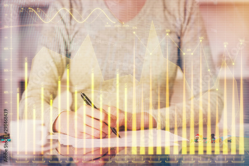 Hand taking notes in notepad. Forex chart holograms in front. Concept of research. Double exposure