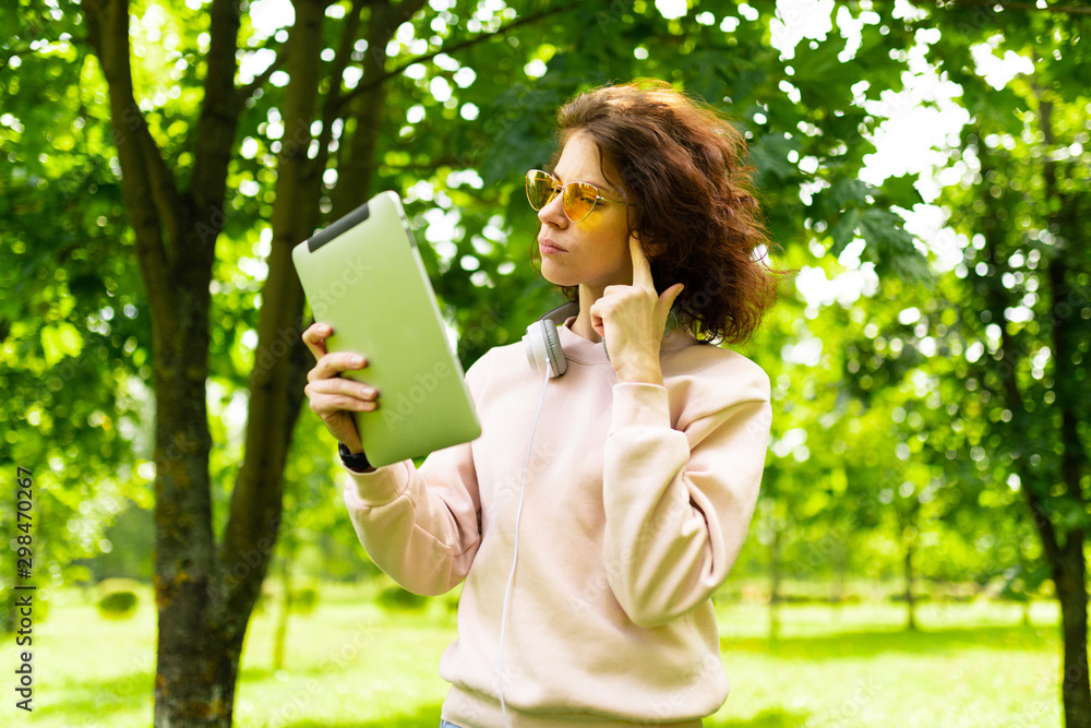 Young caucasian woman with perfect smile, plump lips, glasses, earphones, walks in nature, holds a tablet in hands and thinks Young caucasian woman with perfect smile, plump lips, glasses, earphones, walks in nature, holds a tablet in hands and thinks