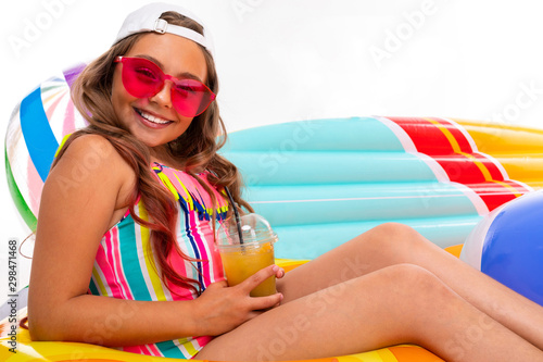 Girl with long crucified hair in sunglasses and swimsuit sits on inflatable mattresses with fresh fruit cocktail isolated on a white background