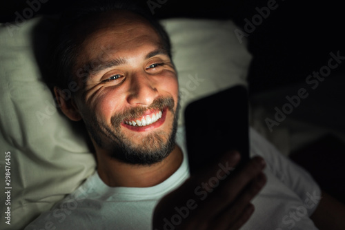 Bearded happy young man is lying in his bed and smiling while watching something on his phone. It is clearly very late at night.