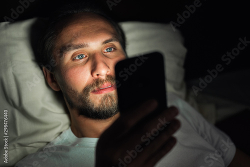 Young handsome and tired man with a beard cannot sleep and is watching something on his phone at night.