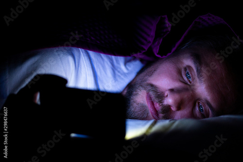 Bearded young man is lying in bed under his blanket. He cannot sleep and is watching something on his mobile phone.