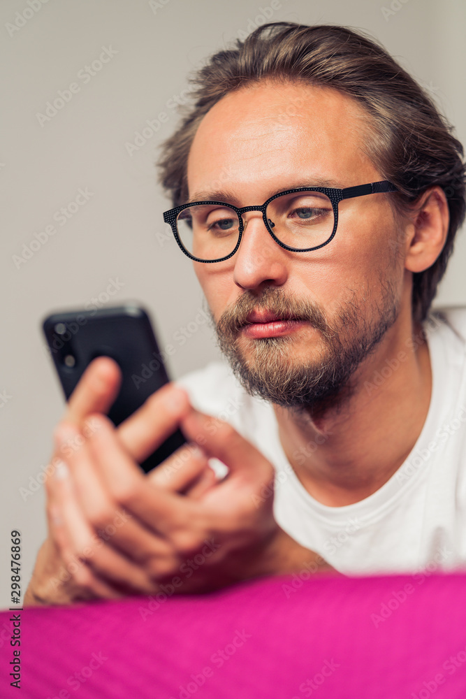 Rather serious young man wearing glasses lying in bed while looking at his mobile phone Rather serious young man wearing glasses lying in bed while looking at his mobile phone