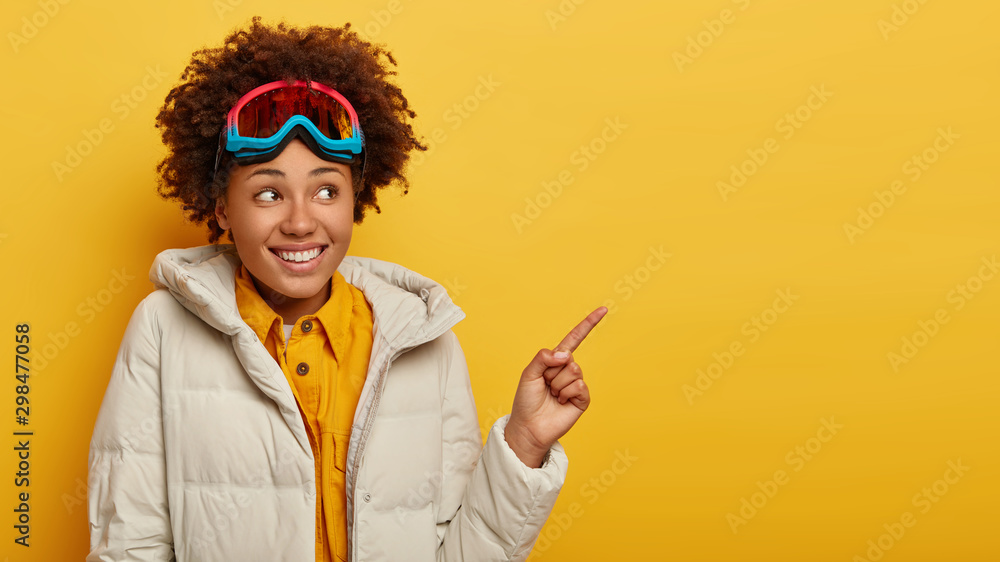 Happy female snowboarder wears skiing mask, looks away and points index finger on blank space, rests in mountains, dressed in warm white jacket, promots something on copy space. Active lifestyle - obrazy, fototapety, plakaty 