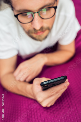 Rather serious young man wearing glasses lying in bed while looking at his mobile phone