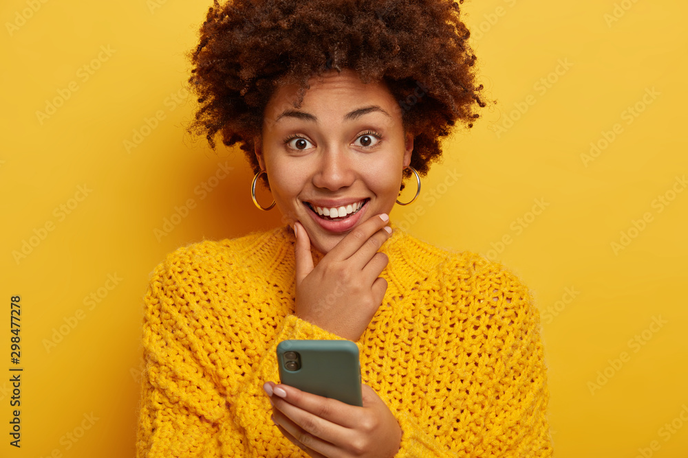 Portrait of good looking smiling female touches chin, smiles broadly, uses cellphone, searches dating application, wears yellow sweater, models indoor. Happy technology mobile user with gadget Portrait of good looking smiling female touches chin, smiles broadly, uses cellphone, searches dating application, wears yellow sweater, models indoor. Happy technology mobile user with gadget