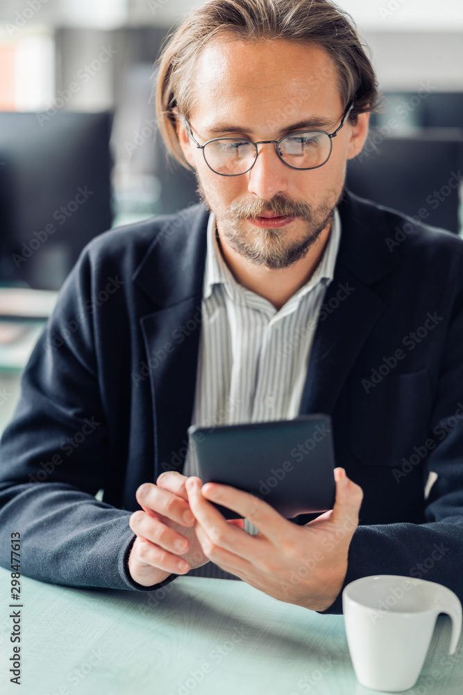 Young handsome man in glasses reading an ebook at work. Coffee cup visible in front of him. Young handsome man in glasses reading an ebook at work. Coffee cup visible in front of him.