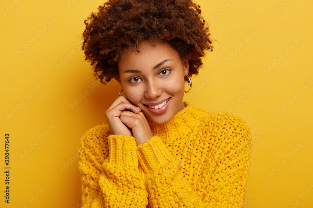 Headshot of adorable romantic woman has pleasant smile, tilts head on hands, has tender look, has dark curly hair, wears cozy winter sweater, isolated over yellow background. Happy emotions. Headshot of adorable romantic woman has pleasant smile, tilts head on hands, has tender look, has dark curly hair, wears cozy winter sweater, isolated over yellow background. Happy emotions.