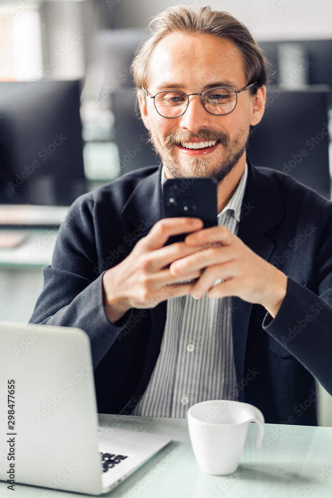 Young handsome man in glasses is interacting with his mobile phone while being at work. Happiness and content at work and being always reachable concepts. Young handsome man in glasses is interacting with his mobile phone while being at work. Happiness and content at work and being always reachable concepts.