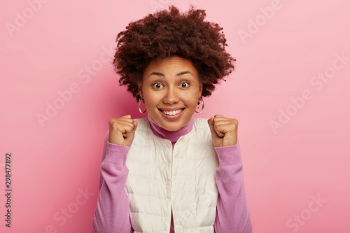 Glad dark skinned woman makes fist bump from happiness, celebrates winning huge bet, wears white vest, casual jumper, smiles positively, enjoys awesome event, makes victory gesture, poses indoor