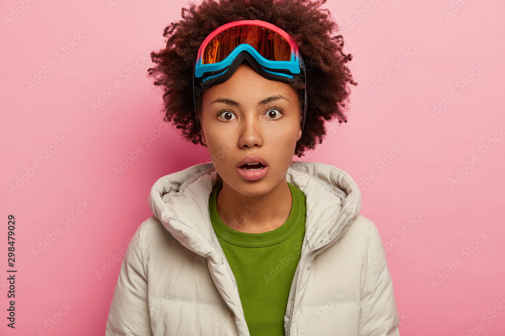 Shocked young woman in winter clothes, spends holidays actively, recreats in mountains, enjoys skiing, wears snowboarding mask on head, holds breath from wonder, isolated over pink background Shocked young woman in winter clothes, spends holidays actively, recreats in mountains, enjoys skiing, wears snowboarding mask on head, holds breath from wonder, isolated over pink background