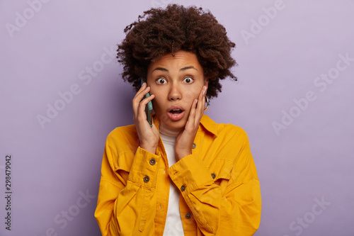 Portrait of attractive Afro woman gets surprising news during telephone call, touches cheek, wears bright jellow jacket, has eyes popped out and mouth opened hears astonishing relevation stands indoor photo