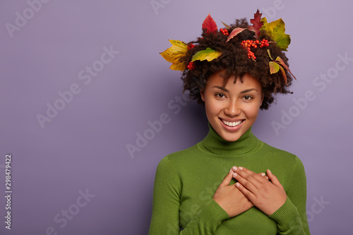 Adorable young Afro American lady feels gratitude, has hands folded on chest, weras long sleeved green jumper, leaves on curly hair, feels great and enjoys day, says thank you for compliment photo