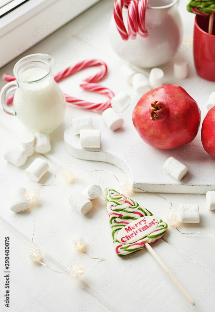 Christmas sweets pomegranate, milk, marshmallows, candies on a white background by the window Christmas sweets pomegranate, milk, marshmallows, candies on a white background by the window