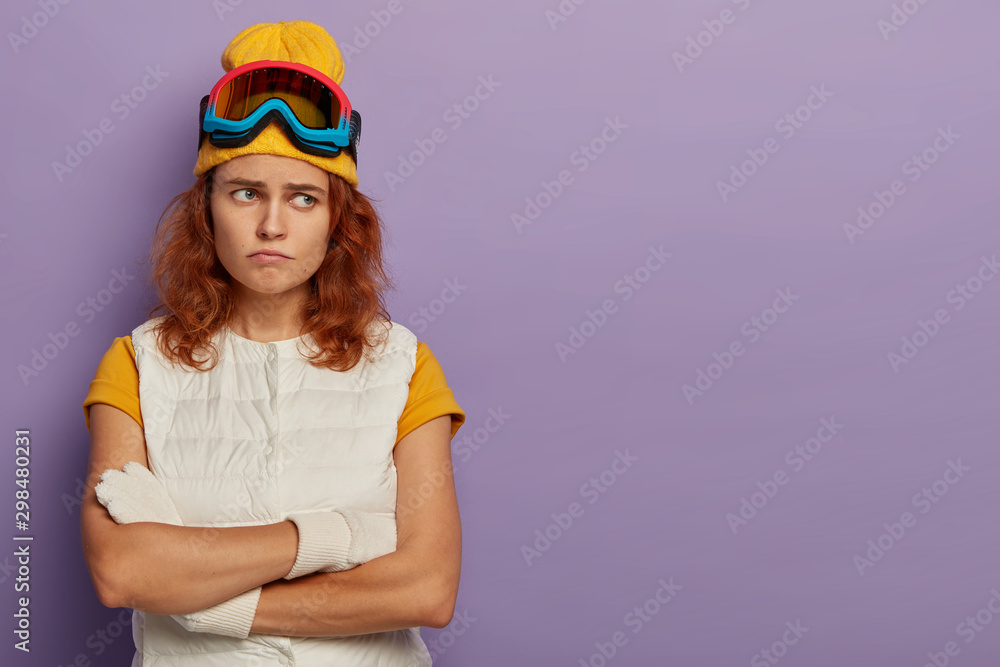 Φωτογραφία Dissatisfied redhaired woman keeps hands crossed, frowns face, being in bad mood after sledding, wears ski goggles and white vest, discontent with something, isolated over purple background