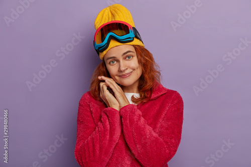Skiing, winter, sport concept. Beautiful woman tilts head, wears yellow hat and red jacket, has fun, enjoys favourite hobby, uses snowboarding glasses looks gladfully at camera isolated on violet wall