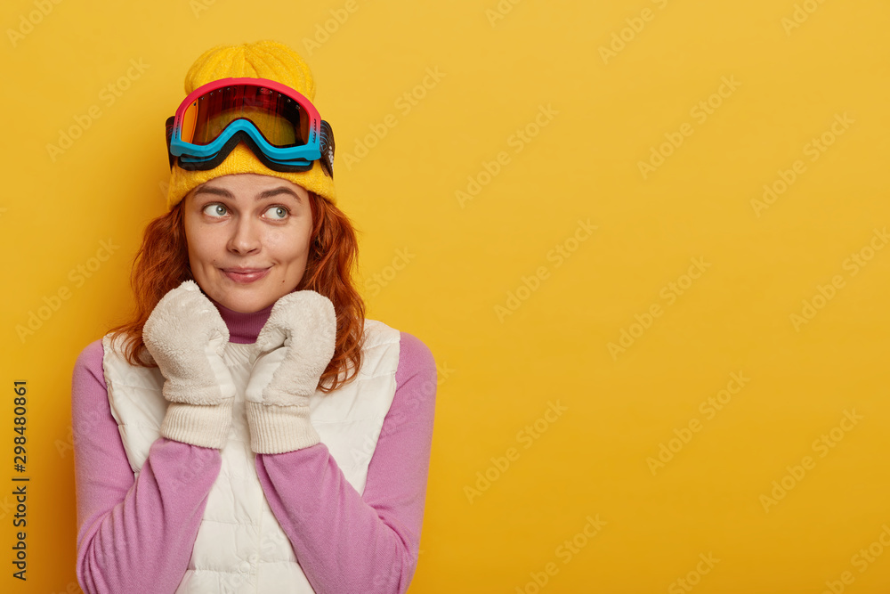 Adorable female tourist has red hair, wears yellow hat with ski goggles on head, white soft mittens, purple jumper with white vest, has active rest during snowy weather, looks away, poses indoor Adorable female tourist has red hair, wears yellow hat with ski goggles on head, white soft mittens, purple jumper with white vest, has active rest during snowy weather, looks away, poses indoor